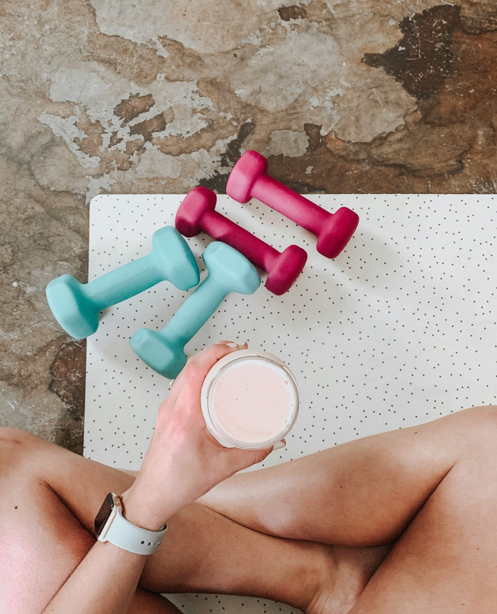 woman sitting with only her legs and an outstretched arm holding a shake showing. Sitting on a yoga mat with a set of light weights in front of her. 