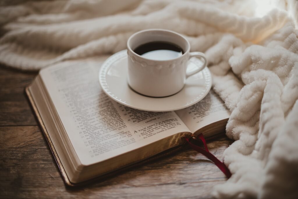 A cup of coffee on a saucer resting on an open book, with a cozy blanket draped nearby.