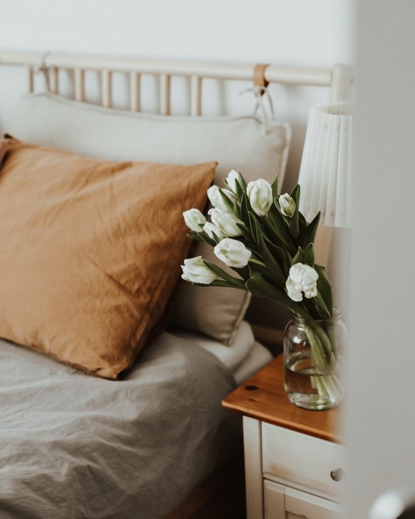 Close-up of a bed with a pillow and part of the comforter visible, a vase of flowers on the nightstand, and a lamp beside it.