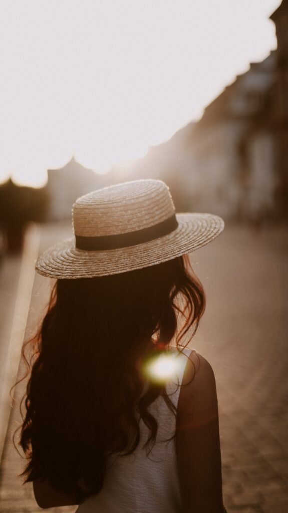 A woman with long hair and a hat, facing away from the camera, with the sun shining brightly in the background.