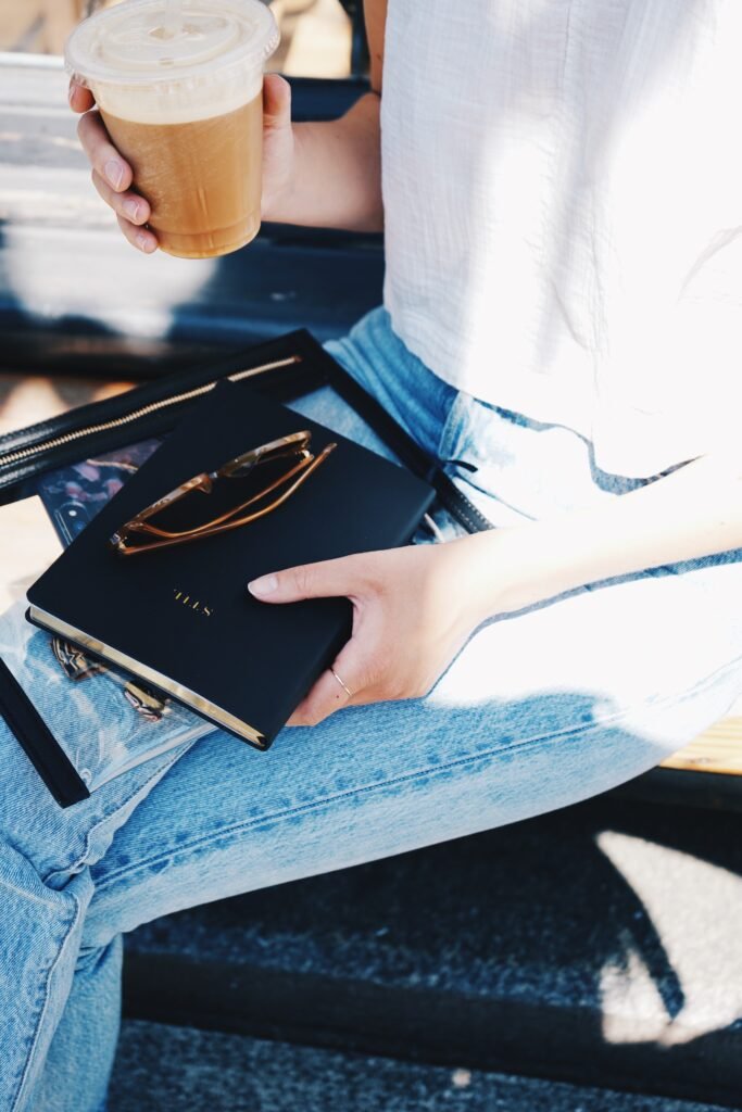 Close up of a Woman sitting with a coffee in one hand and holding her planner and glasses in the other hand balanced on her lap.