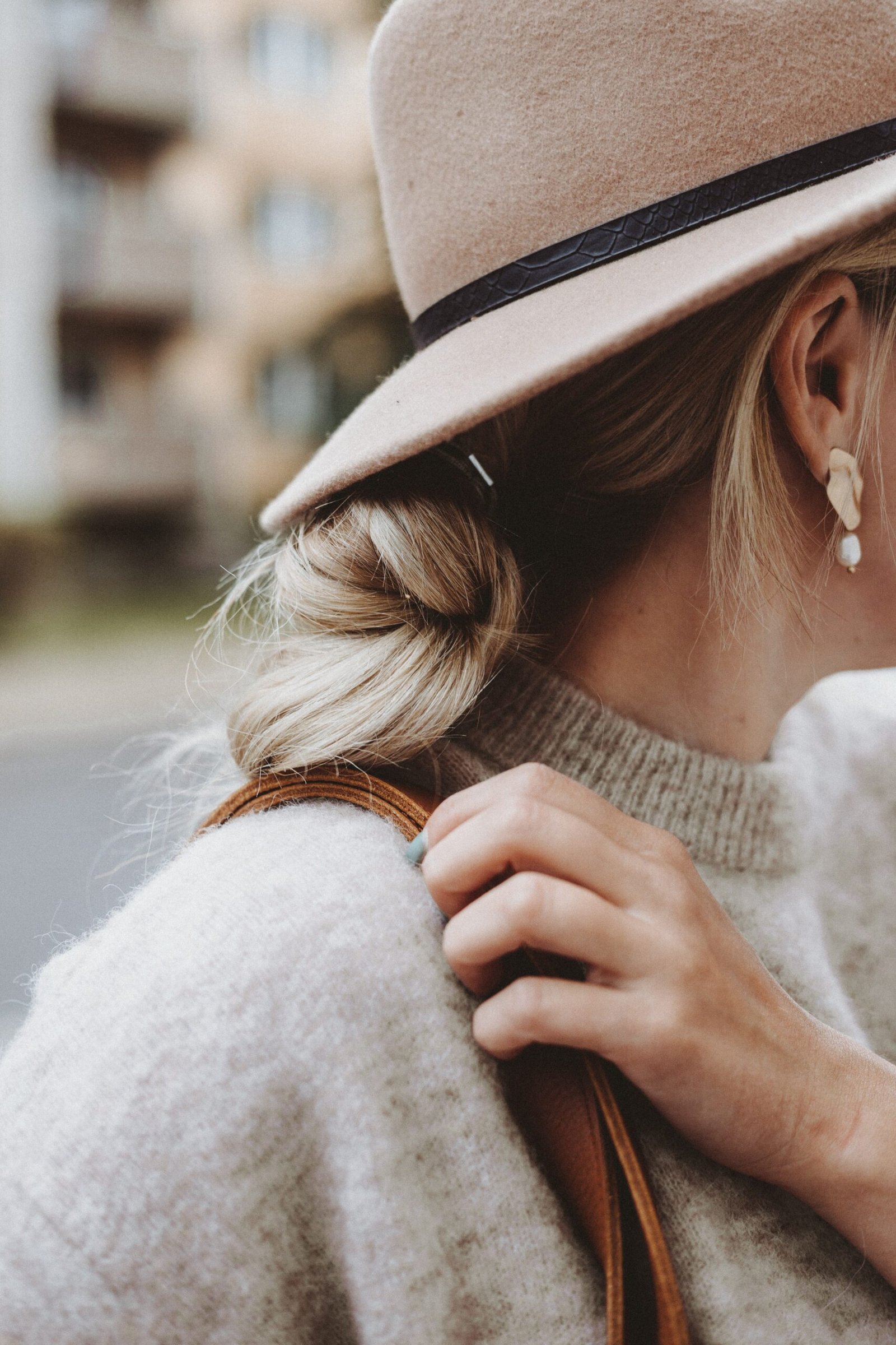 A woman with a bun wears a hat and turns her head to reveal her earrings. She clutches the strap of her purse in this close-up shot from her shoulders up.